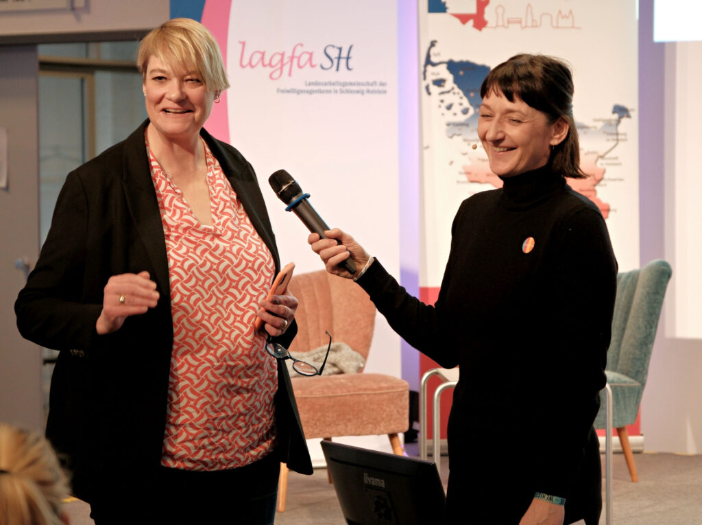 Zwei lachende Frauen, eine hält der anderen ein Mikro hin